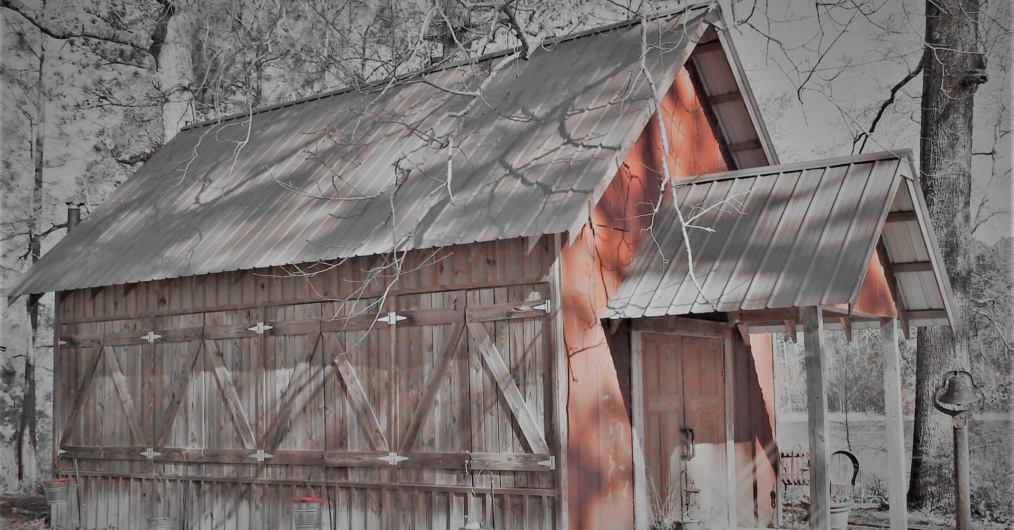 A weathered wooden barn with cross-braced double doors and a small gabled entry porch among bare winter trees, with a lake behind and the gable tinted red against a nearly black-and-white scene